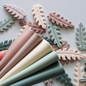 Può includere: Un set di sei forme di foglie in pasta polimerica in tonalità di verde, marrone e rosa. Le foglie sono disposte a ventaglio, con le punte delle foglie rivolte verso la parte superiore dell'immagine.