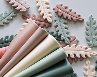 May include: A set of six polymer clay leaf shapes in shades of green, brown, and pink. The leaves are arranged in a fan shape, with the tips of the leaves pointing towards the top of the image.