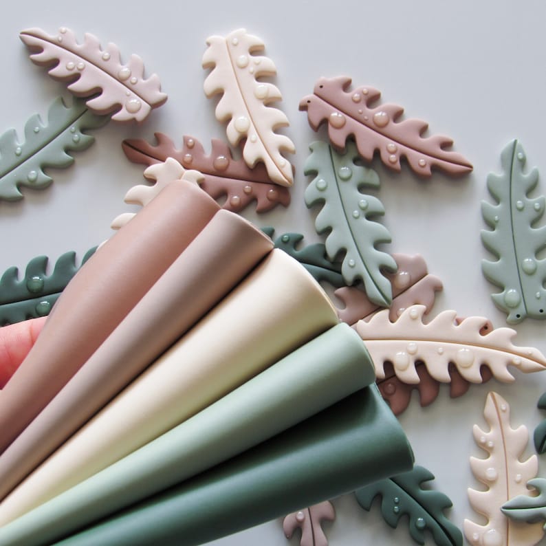 May include: A collection of smooth, matte-finish fern leaf shapes in shades of sage green, cream, beige, and dusty rose, scattered alongside a fan-shaped stack of solid-colored sheets in matching muted tones of green, beige, and brown.