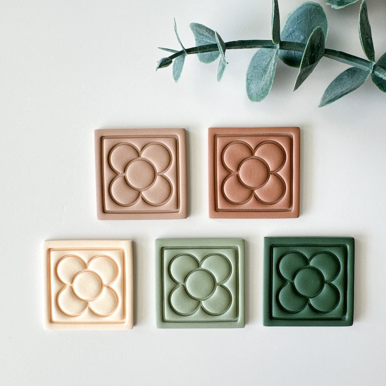 May include: Five square tiles arranged in two rows, each featuring a raised embossed flower design with a central circle and four surrounding petals. The tiles are colored in shades of light beige, terracotta, tan, sage green, and dark forest green.