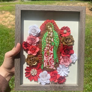 May include: A framed artwork featuring a central image of a religious figure, surrounded by red, gold, and white paper flowers. The figure is depicted in green and pink attire, with a gold necklace. The frame is a light gray color.