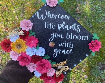 Può includere: Un cappello di laurea nero decorato con fiori di carta in tonalità rosa, rosso e bianco. Il cappello ha la citazione "Wherever life plants you, bloom with grace" scritta in bianco.