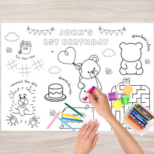 Op de afbeelding: Een witte activiteit placemat voor een eerste verjaardag, met een berenthema en de tekst "John's 1st Birthday". Inclusief verbind-de-stippen, tic-tac-toe, een doolhof en een taart om te versieren. Kleurpotloden en stiften zijn inbegrepen.