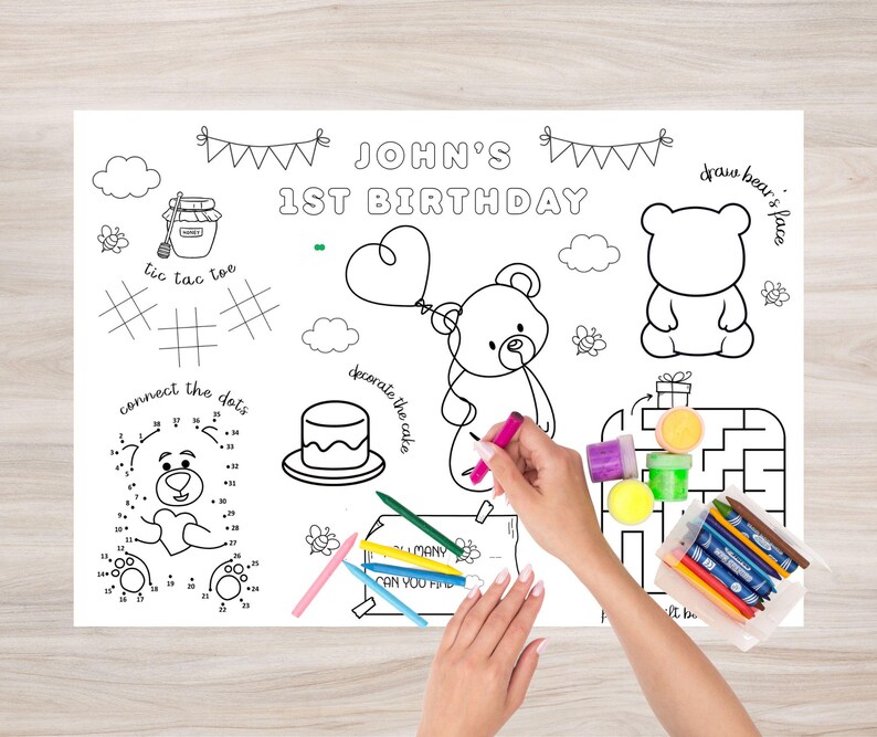 Op de afbeelding: Een witte activiteit placemat voor een eerste verjaardag, met een berenthema en de tekst "John's 1st Birthday". Inclusief verbind-de-stippen, tic-tac-toe, een doolhof en een taart om te versieren. Kleurpotloden en stiften zijn inbegrepen.