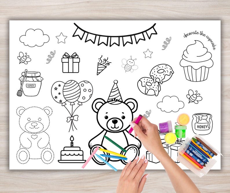 Op de afbeelding: Een witte kleurplaat met illustraties met een verjaardagsthema, waaronder een teddybeer, taart, ballonnen en cupcakes. De tekst "decorate the cupcake" is zichtbaar. Een hand houdt een roze kleurpotlood vast, klaar om te kleuren.