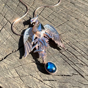 May include: A copper-colored pendant necklace featuring a stylized phoenix with outstretched wings and a dark blue, round gemstone. The necklace is displayed on a weathered wooden surface, with a small wooden box in the background.