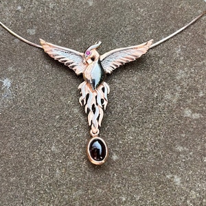 Könnte beinhalten: Eine Halskette aus Silber und Gold mit einem Phönix-Anhänger. Der Phönix ist mit schwarzem Email verziert und hat ein rotes Edelsteinauge. Der Anhänger ist an einer silbernen Kette mit einem schwarzen Edelsteintropfen befestigt.