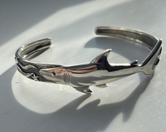 May include: A silver cuff bracelet featuring a detailed shark design. The open-ended bracelet has a shark figure with a dorsal fin, tail, and gills. The shark is swimming through waves with a star accent. The bracelet is made of a shiny metal.