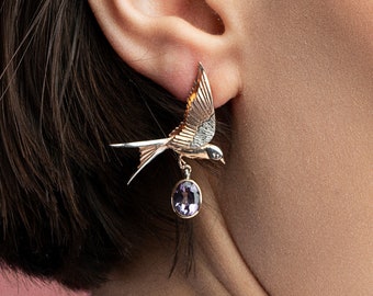 May include: A pair of gold earrings featuring a detailed bird design with a dangling amethyst gemstone. The earrings are worn by a person with dark hair and a neutral makeup look.