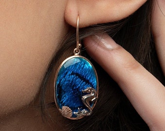May include: Gold tone earrings with a blue oval-shaped stone featuring a seahorse and seashells.