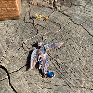 May include: A detailed pendant necklace featuring a phoenix design. The pendant is made of copper-colored metal with blue and silver accents. A round, dark blue stone hangs below the phoenix. The necklace is displayed on a weathered wooden surface.