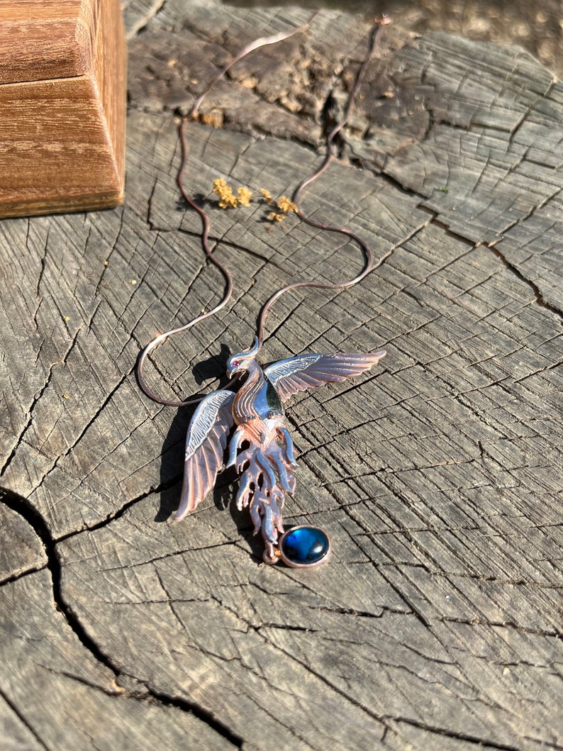 May include: A detailed pendant necklace featuring a phoenix design. The pendant is made of copper-colored metal with blue and silver accents. A round, dark blue stone hangs below the phoenix. The necklace is displayed on a weathered wooden surface.
