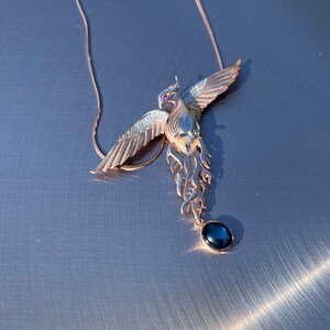 May include: A rose gold phoenix pendant necklace with a dark blue gemstone. The phoenix has outstretched wings and a red gemstone eye. The necklace is on a silver surface.