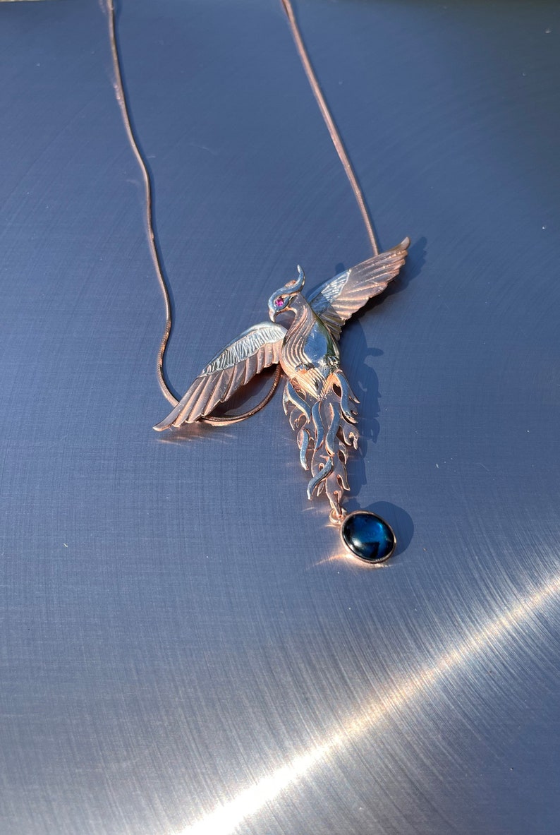 May include: A rose gold phoenix pendant necklace with a dark blue gemstone. The phoenix has outstretched wings and a red gemstone eye. The necklace is on a silver surface.