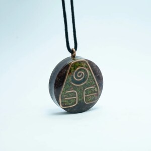 May include: A round pendant necklace featuring a copper-coloured symbol of a triangle with a spiral inside. The pendant is crafted from a dark brown stone with green and red flecks. The necklace is made of black cord.