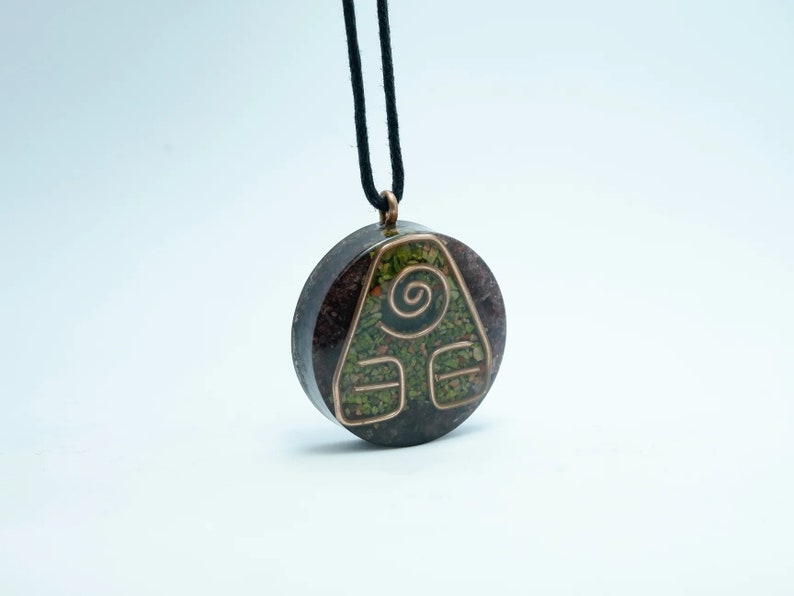May include: A round pendant necklace featuring a copper-coloured symbol of a triangle with a spiral inside. The pendant is crafted from a dark brown stone with green and red flecks. The necklace is made of black cord.