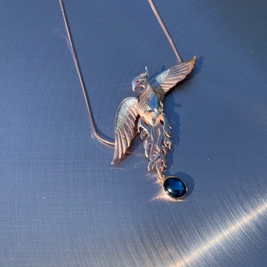 May include: A rose gold phoenix pendant necklace with a dark blue gemstone. The phoenix has outstretched wings and a small red gemstone eye. The necklace chain is delicate and rose gold. Jewelry.