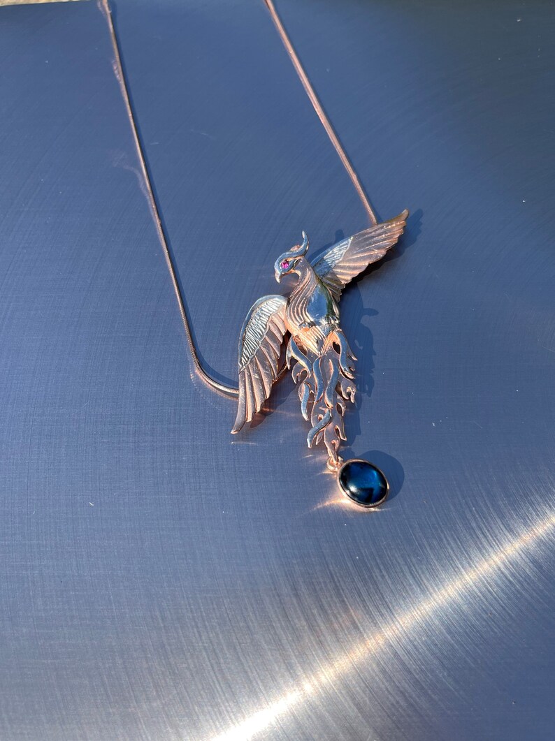 May include: A rose gold phoenix pendant necklace with a dark blue gemstone. The phoenix has outstretched wings and a small red gemstone eye. The necklace chain is delicate and rose gold. Jewelry.