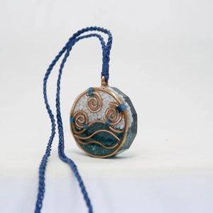 May include: A round pendant necklace with a blue and white design. The pendant features a stylised wave design with copper wire accents. The necklace is made with a blue cord.