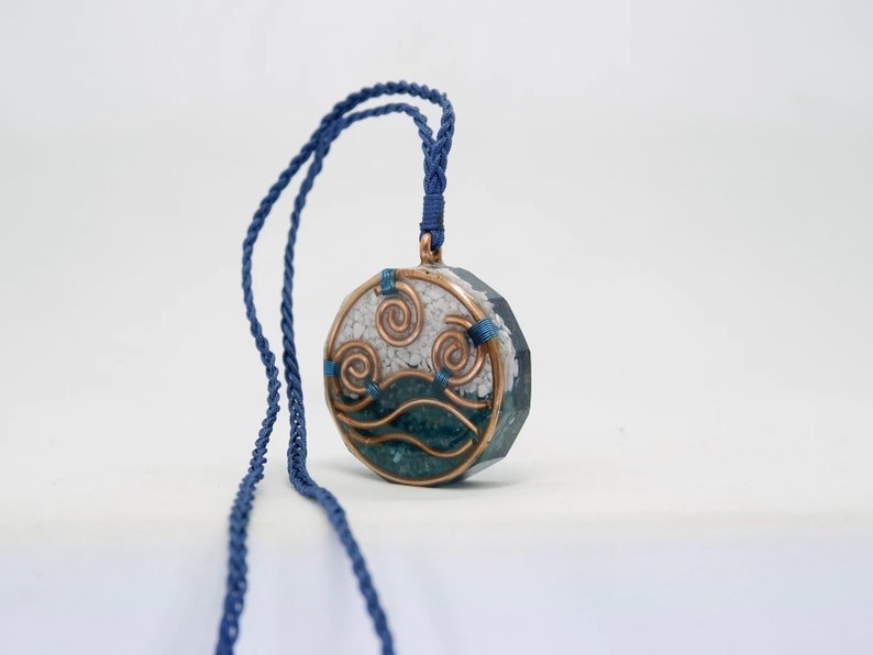 May include: A round pendant necklace with a blue and white design. The pendant features a stylised wave design with copper wire accents. The necklace is made with a blue cord.