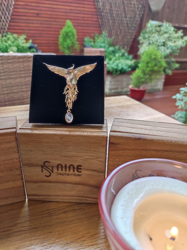 May include: A gold phoenix pendant with a moonstone drop, displayed on a black velvet cushion. The cushion is on a wooden box with the text "Nine September" engraved on the front.