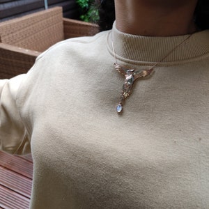 May include: A silver necklace with a bird pendant and a blue gemstone. The bird is in flight with its wings spread wide. The necklace is worn by a person wearing a beige sweater.