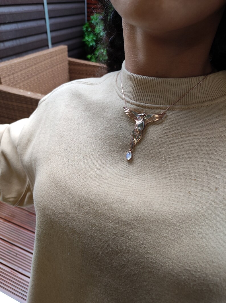 May include: A silver necklace with a bird pendant and a blue gemstone. The bird is in flight with its wings spread wide. The necklace is worn by a person wearing a beige sweater.
