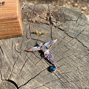 May include: A copper-toned phoenix pendant with a blue gemstone, resting on a weathered wooden surface. The bird's wings are detailed with white and silver accents. A wooden box is partially visible in the background.
