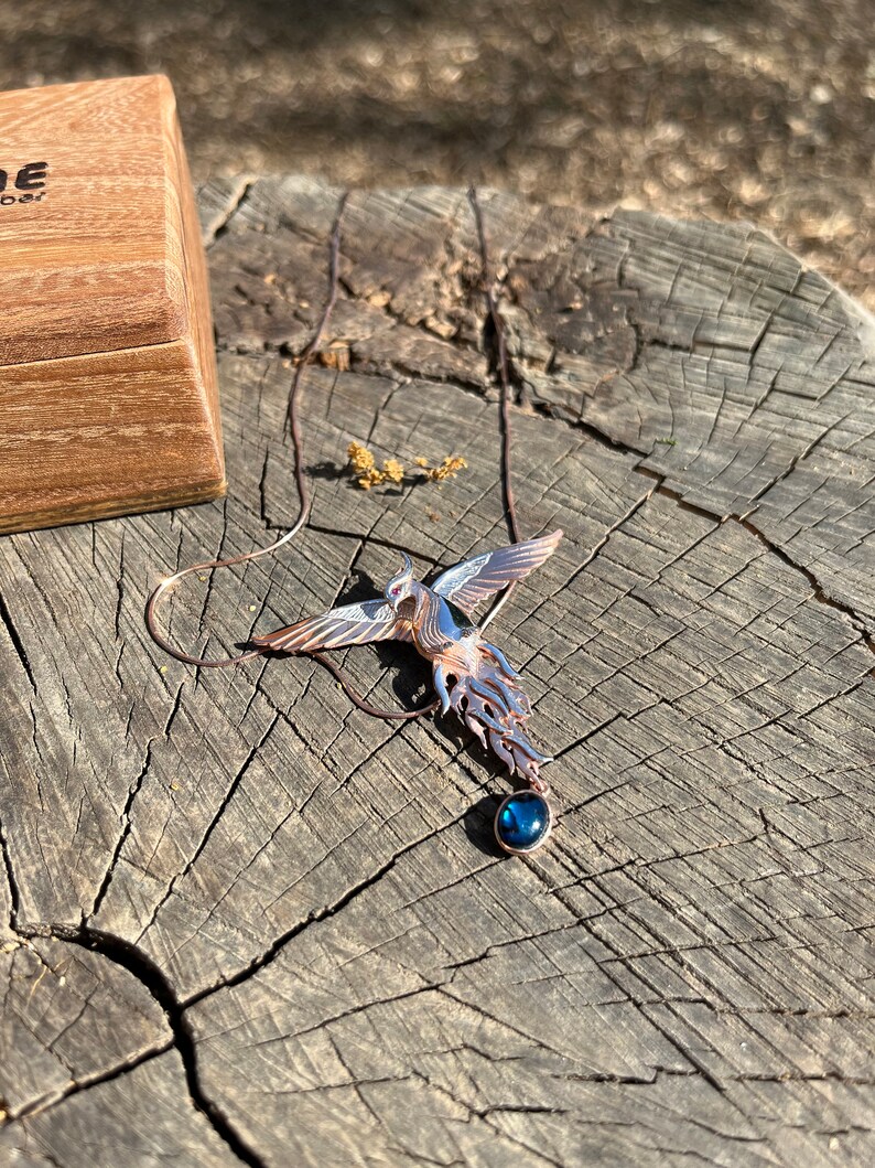 May include: A copper-toned phoenix pendant with a blue gemstone, resting on a weathered wooden surface. The bird's wings are detailed with white and silver accents. A wooden box is partially visible in the background.