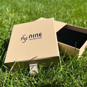 May include: An open, tan-colored box with a black interior, resting on green grass. The box has a textured surface and the words "S Nine September" printed on the lid in black. A light pink ribbon is attached to the box.