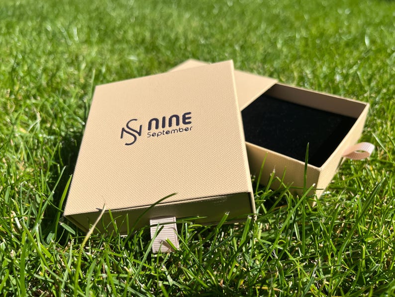 May include: An open, tan-colored box with a black interior, resting on green grass. The box has a textured surface and the words "S Nine September" printed on the lid in black. A light pink ribbon is attached to the box.