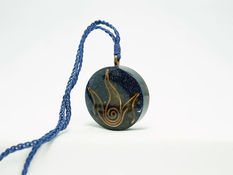 May include: A blue and gold necklace with a round pendant featuring a stylised flame design. The pendant is made of resin and has a glittery finish.