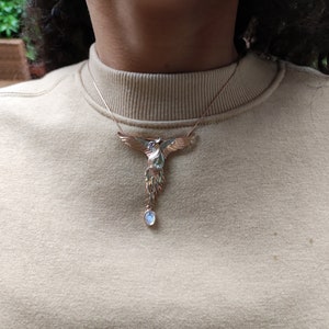 May include: A gold-toned necklace with a detailed phoenix pendant and a single, round, light blue gemstone hanging from the bottom.