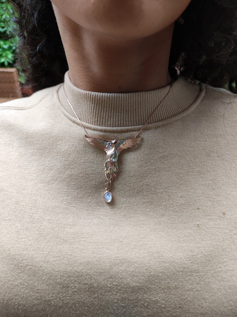 May include: A gold-toned necklace with a detailed phoenix pendant and a single, round, light blue gemstone hanging from the bottom.