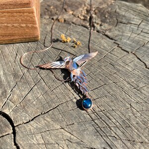 May include: A copper-toned phoenix pendant with outstretched wings and a blue gemstone. The bird-shaped necklace is displayed on a weathered wooden surface, with a wooden box in the background.