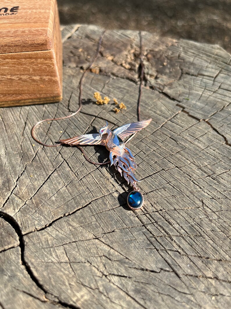 May include: A copper-toned phoenix pendant with outstretched wings and a blue gemstone. The bird-shaped necklace is displayed on a weathered wooden surface, with a wooden box in the background.