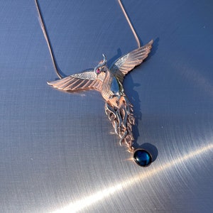 May include: A rose gold phoenix pendant necklace. The bird has outstretched wings, a red gemstone eye, and a dangling dark blue teardrop-shaped stone. The necklace is displayed on a metallic surface.
