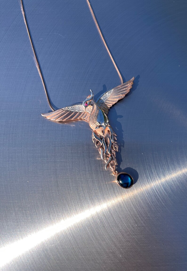 May include: A rose gold phoenix pendant necklace. The bird has outstretched wings, a red gemstone eye, and a dangling dark blue teardrop-shaped stone. The necklace is displayed on a metallic surface.