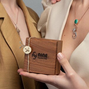 May include: A small, rectangular wooden box with the text "S Nine September" is held by a person. The box is tied with twine and a button. A necklace with a pendant is visible.