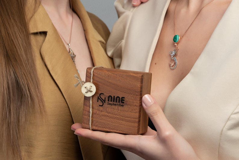 May include: A small, rectangular wooden box with the text "S Nine September" is held by a person. The box is tied with twine and a button. A necklace with a pendant is visible.