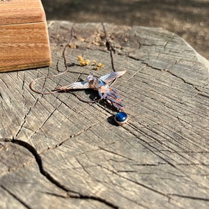 May include: A detailed pendant necklace featuring a stylized bird design with outstretched wings. The pendant has blue and copper tones, with a dark blue gemstone at the bottom. The necklace is displayed on a weathered wooden surface, with a wooden box in the background.