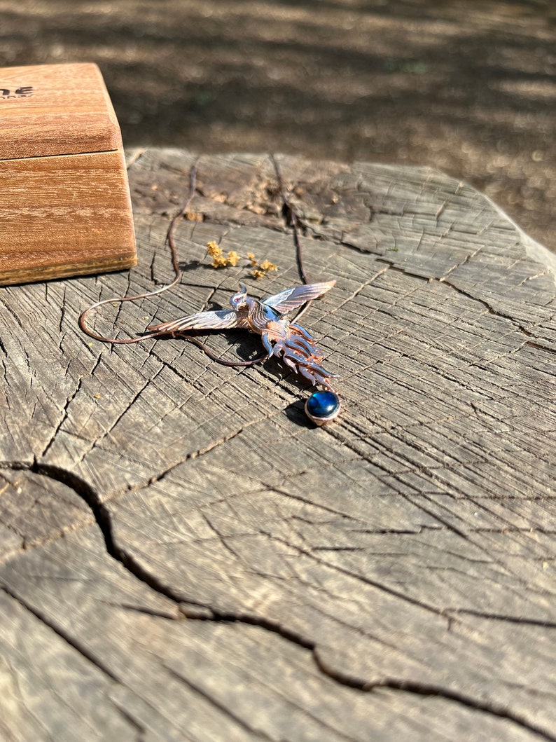 May include: A detailed pendant necklace featuring a stylized bird design with outstretched wings. The pendant has blue and copper tones, with a dark blue gemstone at the bottom. The necklace is displayed on a weathered wooden surface, with a wooden box in the background.