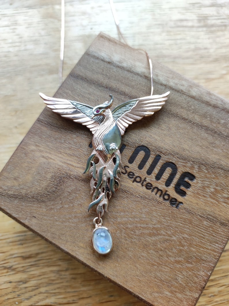 May include: A gold and silver phoenix pendant necklace with a red gemstone eye and a moonstone drop. The necklace is displayed in a wooden box with the inscription "nine September".