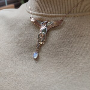 May include: A gold and silver phoenix necklace with a moonstone pendant. The phoenix is detailed with red eyes and a spread wing design.