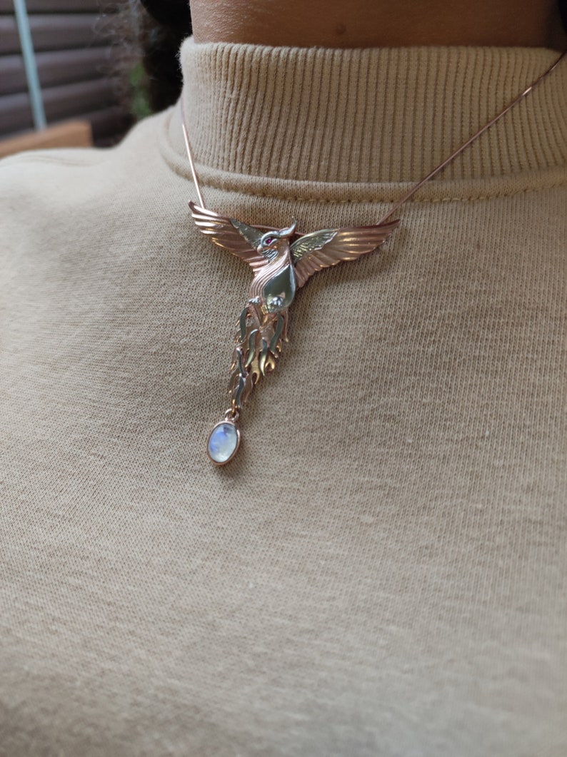 May include: A gold and silver phoenix necklace with a moonstone pendant. The phoenix is detailed with red eyes and a spread wing design.
