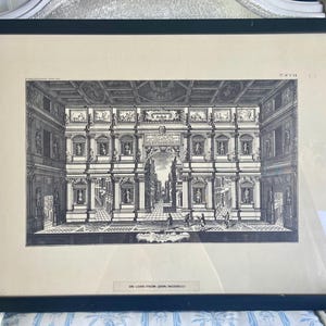 May include: A framed black and white architectural print. The print depicts a detailed interior scene with columns, statues, and figures. The artwork is framed in a black frame and is a classic piece of art.