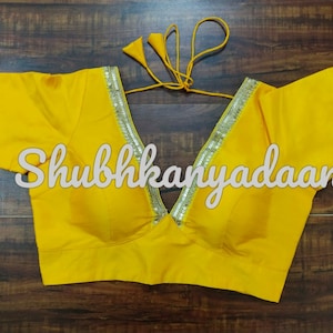 May include: A yellow sleeveless blouse with a deep V-neckline and silver sequin trim. The blouse has a tie closure at the back and the text "Shubhkanyadaan" is printed on the front.
