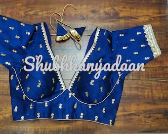 Op de afbeelding: Een blauwe zijden blouse met gouden borduurwerk en kantversiering. De blouse heeft een diepe V-hals en korte mouwen. De tekst "Shubhkanyadaan" is zichtbaar op de voorkant van de blouse.