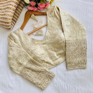 May include: A cream-coloured embroidered blouse with long sleeves and a scoop neckline. The sleeves have a floral pattern and are embellished with sequins.
