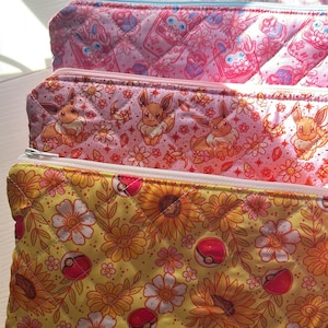Könnte beinhalten: Drei gesteppte Federmäppchen mit Reißverschlüssen. Das obere Etui ist rosa mit einem sich wiederholenden Muster aus Zeichentrickfiguren und Blumen. Das mittlere Etui ist rosa mit Evoli-Figuren und Blumen. Das untere Etui ist gelb mit Sonnenblumen und rot-weißen Pokeball-Designs.